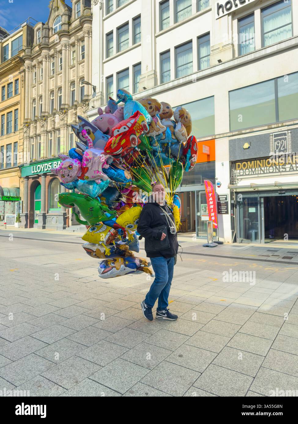 Un venditore di mongolfiere attraversa Queen Street, Cardiff, un caleidoscopio di amati personaggi dei cartoni animati che lo sorvolano nell'aria mattutina. Foto Stock