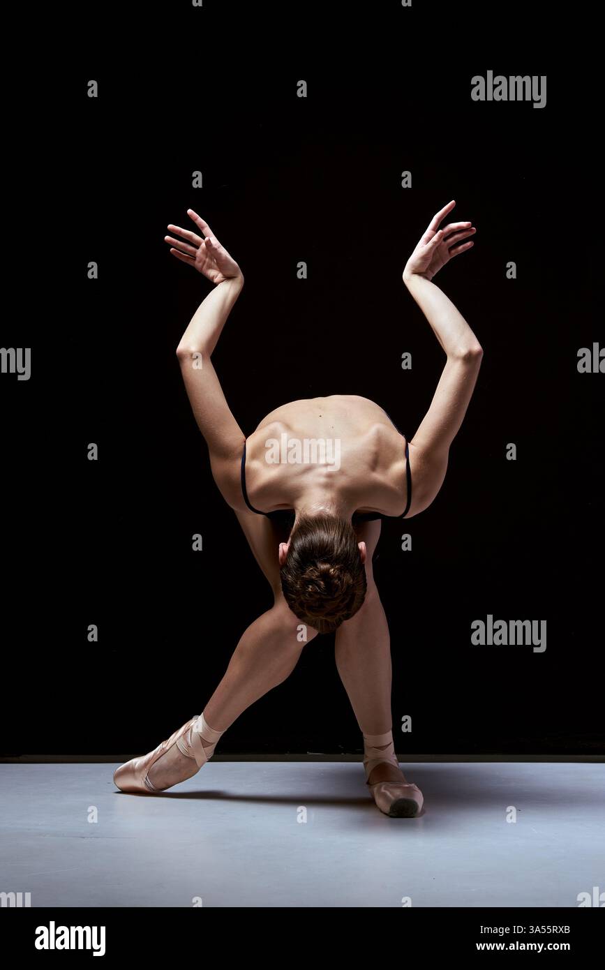 Graziosa ballerina che esegue una posa espressiva con le braccia alzate. Una luce intensa mette in risalto i muscoli della schiena e i movimenti eleganti. Foto Stock