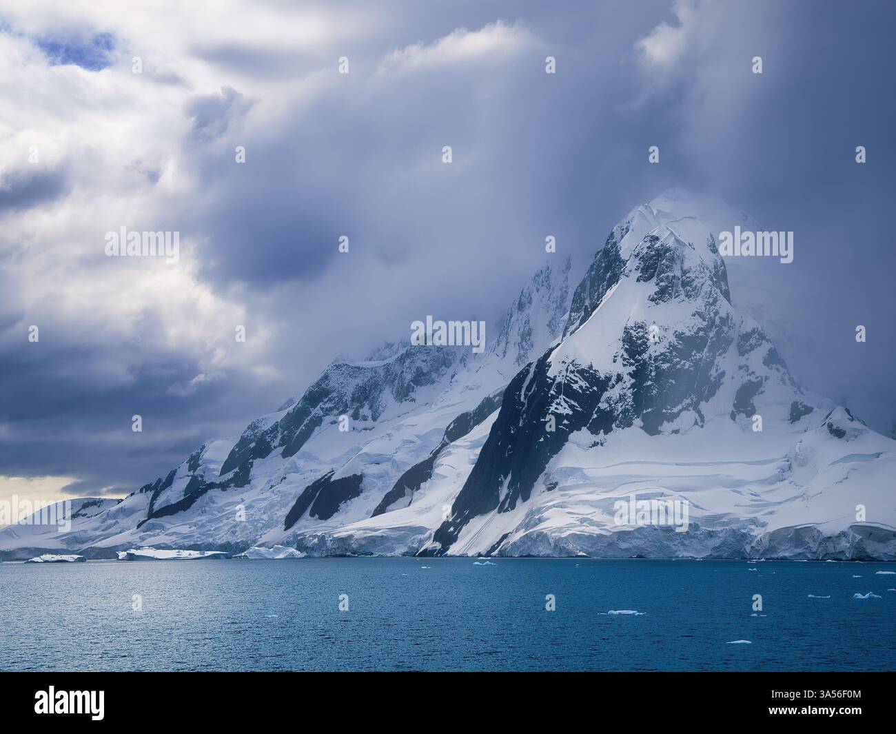 Una delle torreggianti montagne innevate che dominano l'Antartico con un bagliore di sole attraverso le nuvole che illuminano il mare. Foto Stock