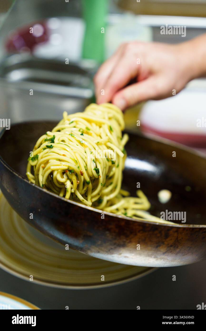 Uno chef prepara con cura una porzione di spaghetti aglio e olio utilizzando pinze in una cucina professionale. La pasta è ricoperta di olio d'oliva, aglio, prezzemolo, Foto Stock