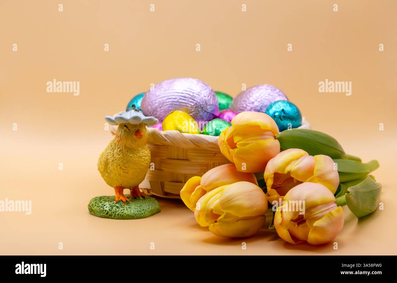 Natura morta di Pasqua con una pulcino decorativa, un cestino di uova di cioccolato colorate e tulipani gialli freschi su un morbido sfondo beige. Foto Stock