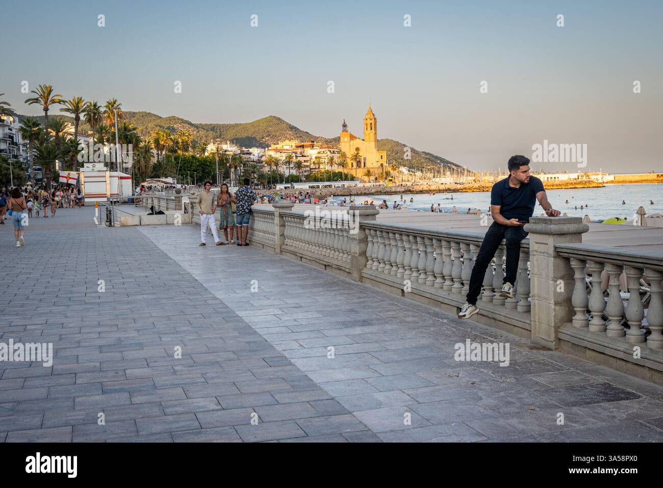 Un uomo parla al telefono sul muro vicino al Passeig Marítim con vista sulla spiaggia e sull'Iglesia de Sant Bartomeu sullo sfondo lontano. Foto Stock