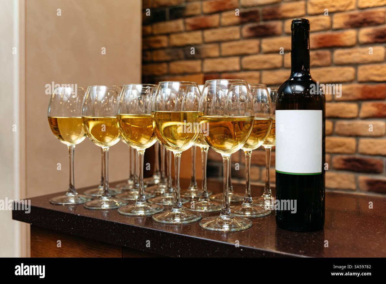 Una fila di eleganti bicchieri da vino riempiti di vino bianco accanto a una bottiglia con un'etichetta vuota, adagiata su una parete di mattoni. Foto Stock