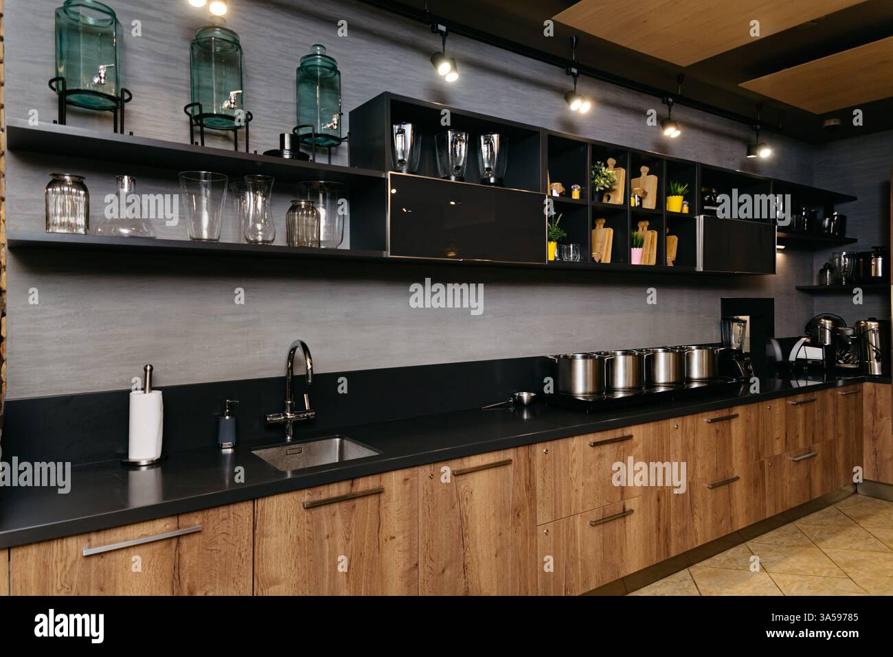 Un'elegante cucina moderna con armadi in legno, ripiani neri, scaffali aperti con vetreria, utensili da cucina e pentole in acciaio inossidabile. Foto Stock