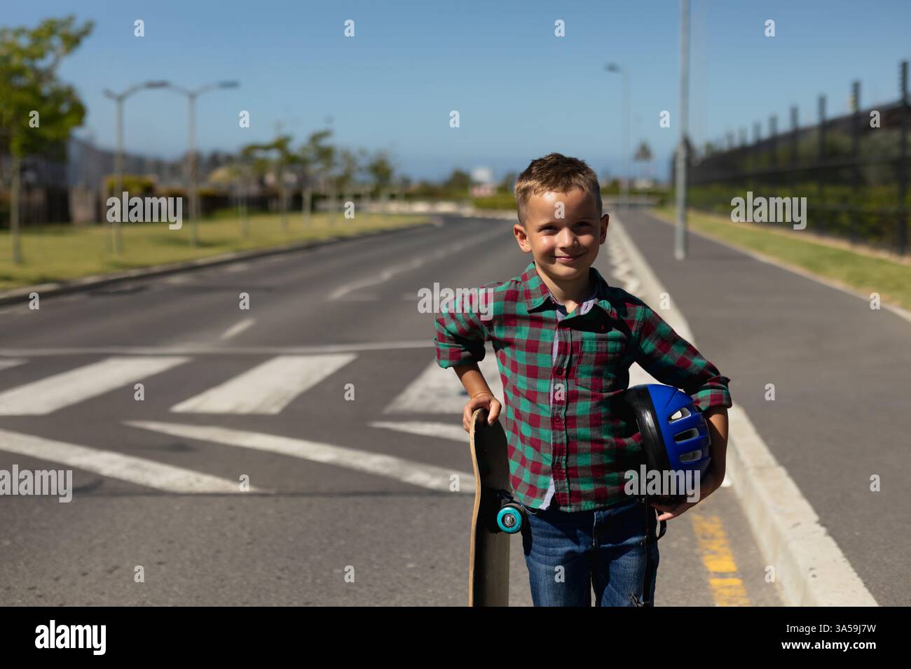 Ragazzo sorridente che tiene in mano skateboard e casco in piedi sulla strada della scuola, copia spazio Foto Stock
