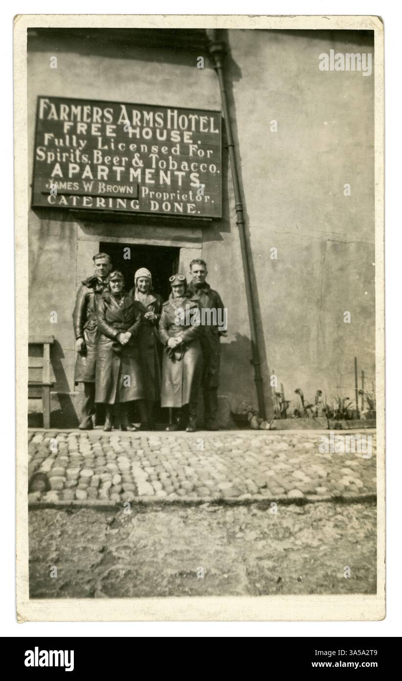 Cartolina originale degli anni '1920/primi anni '1930 di motociclisti primitivi, uomini e donne, su un rideout, indossando trench lunghi in pelle, occhiali, guanti, berretti. Donne moderne! Nel Regno Unito del 1920, le donne ottennero una maggiore indipendenza, compresi i diritti di voto e l'accesso alle professioni, e abbracciarono nuove libertà nella moda, nei comportamenti e nella vita sociale come si vede con queste donne emacipate, una di loro è orgogliosa anche di fumare. Il gruppo di motociclisti sembra che siano stati fuori per un giro in campagna e si trovino fuori dal Farmers Arms Hotel, (James W. Brown è il proprietario) da qualche parte nel Regno Unito Foto Stock