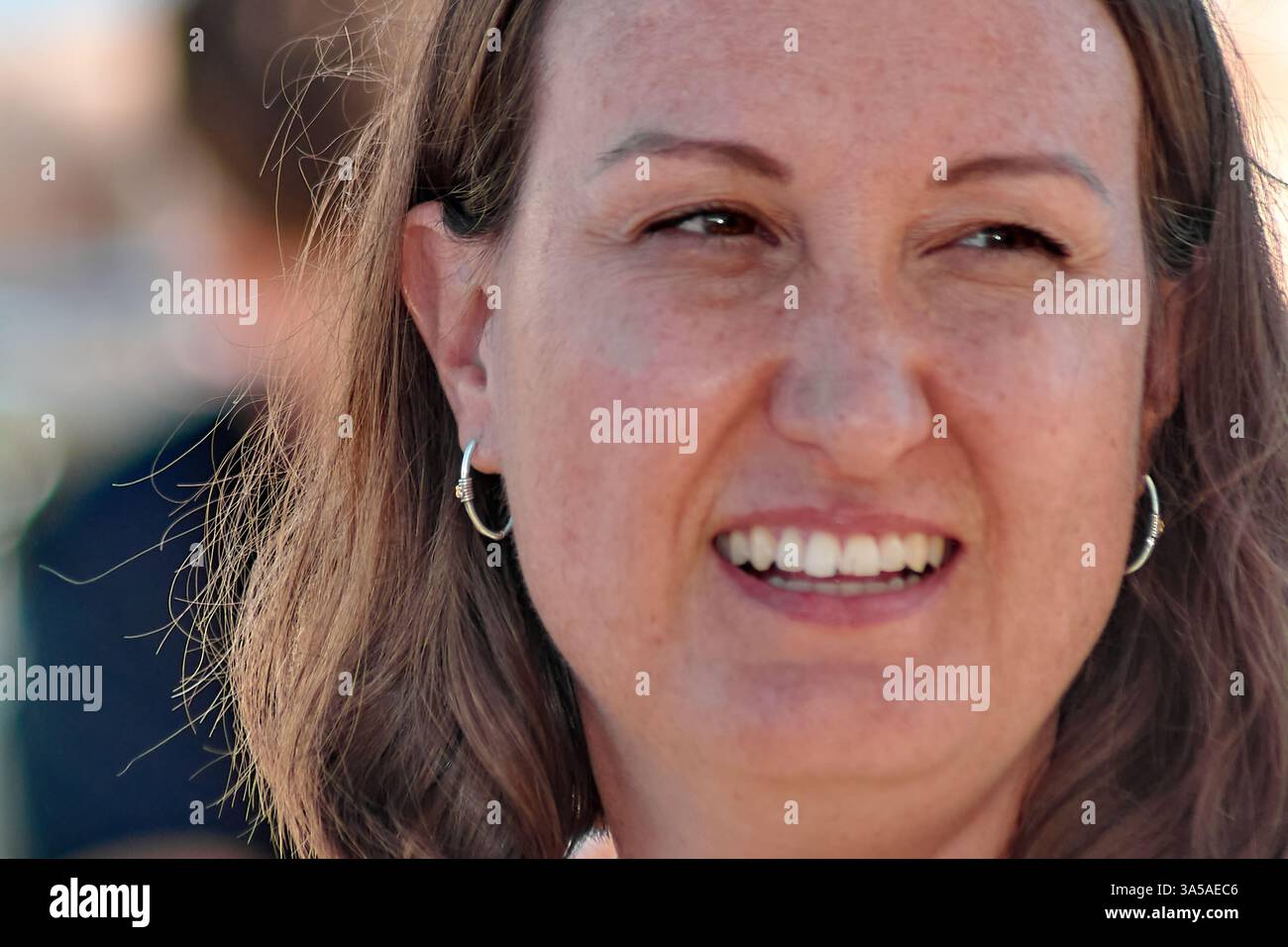 Una donna con un sorriso caldo e invitante, che indossa un top floreale e orecchini a cerchio dorati, catturati alla luce naturale del sole. Foto Stock