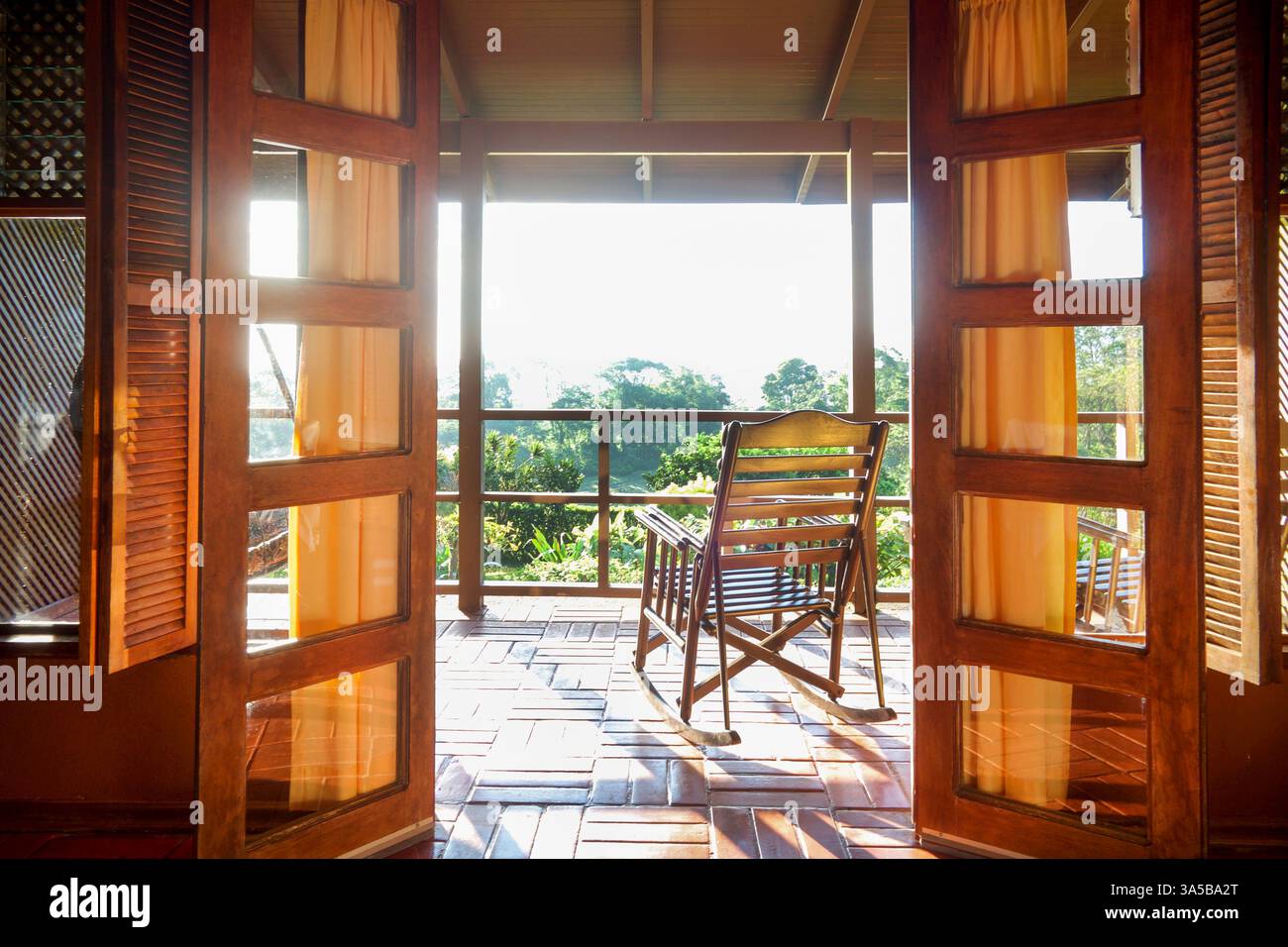 due splendide porte in vetro di legno che si aprono su un balcone con una bella sedia a dondolo di legno che si affaccia su un giardino tropicale con sole luminoso Foto Stock