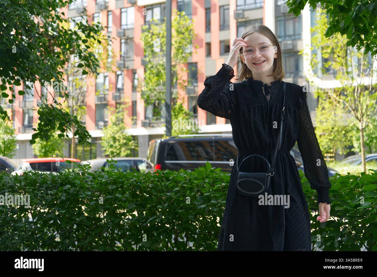 Una ragazza sorride all'esterno, circondata da vegetazione lussureggiante e da edifici cittadini sullo sfondo. Foto Stock