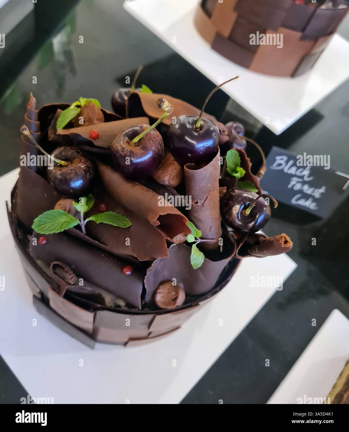 Una torta della foresta nera splendidamente lavorata presenta ricci di cioccolato fondente, ciliegie fresche e foglie di menta, esposti in una panetteria locale. Foto Stock