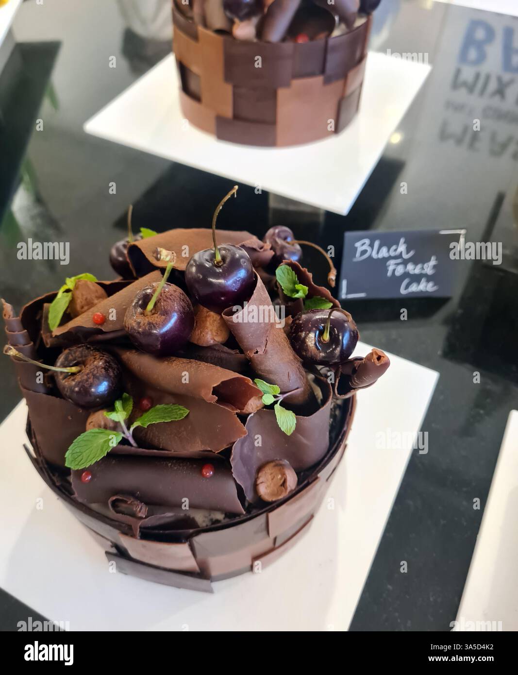 Una torta della foresta nera splendidamente lavorata presenta ricci di cioccolato fondente, ciliegie fresche e foglie di menta, esposti in una panetteria locale. Foto Stock