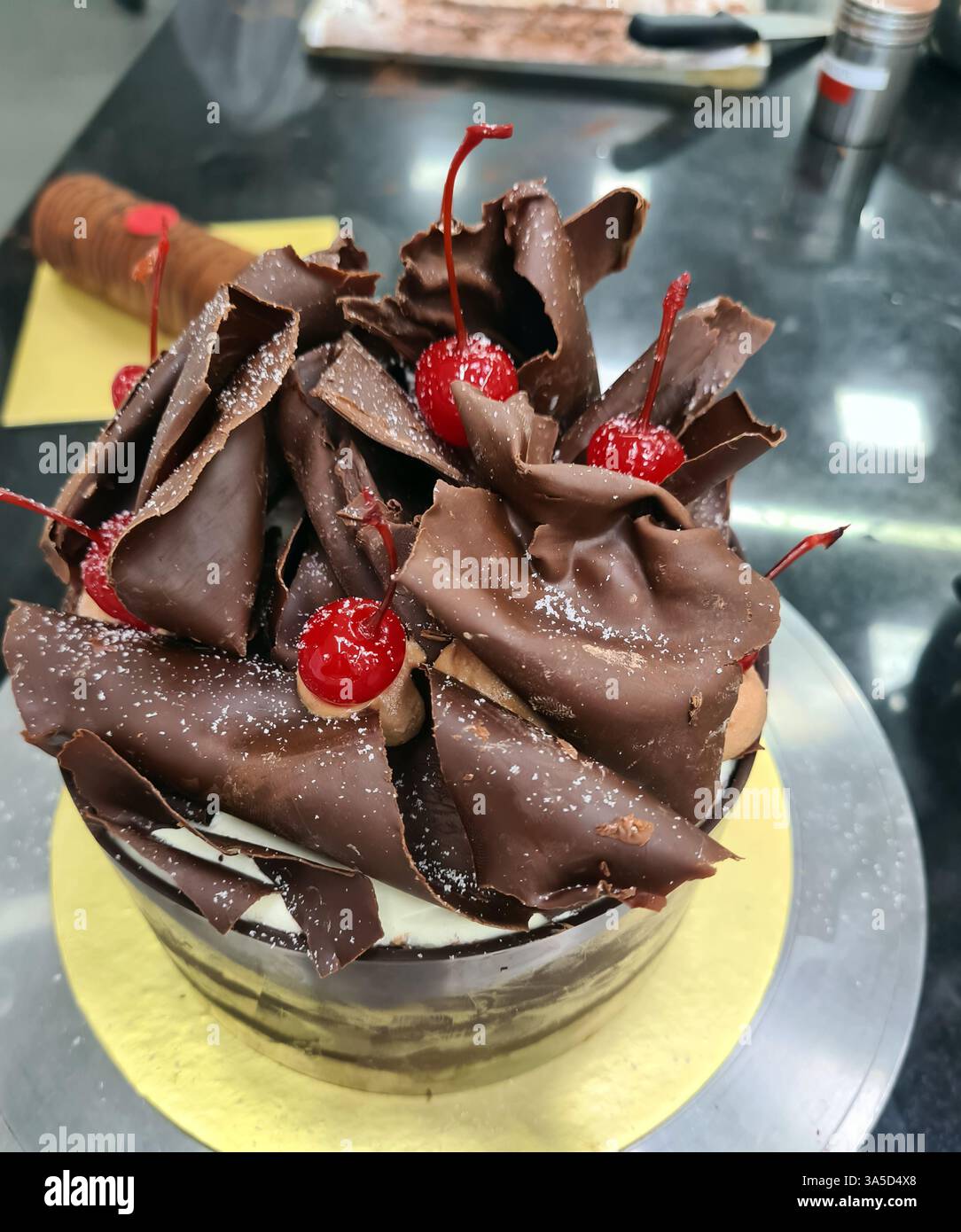 Una torta al cioccolato splendidamente lavorata presenta strati di delicati scaglie di cioccolato e ciliegie brillanti, attirando i clienti in una panetteria affollata. Foto Stock