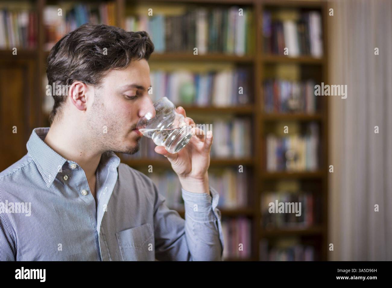 Giovane in camicia bevendo acqua da cristallo sullo sfondo di scaffali Foto Stock Giovane in camicia bevendo acqua da cristallo sullo sfondo di scaffali Foto Stock