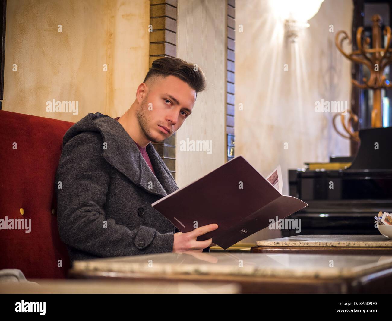 Giovane uomo alla moda seduto accanto al tavolo e con un menu' di lettura nel Cafe', indossando cappotto e vestiti invernali Foto Stock