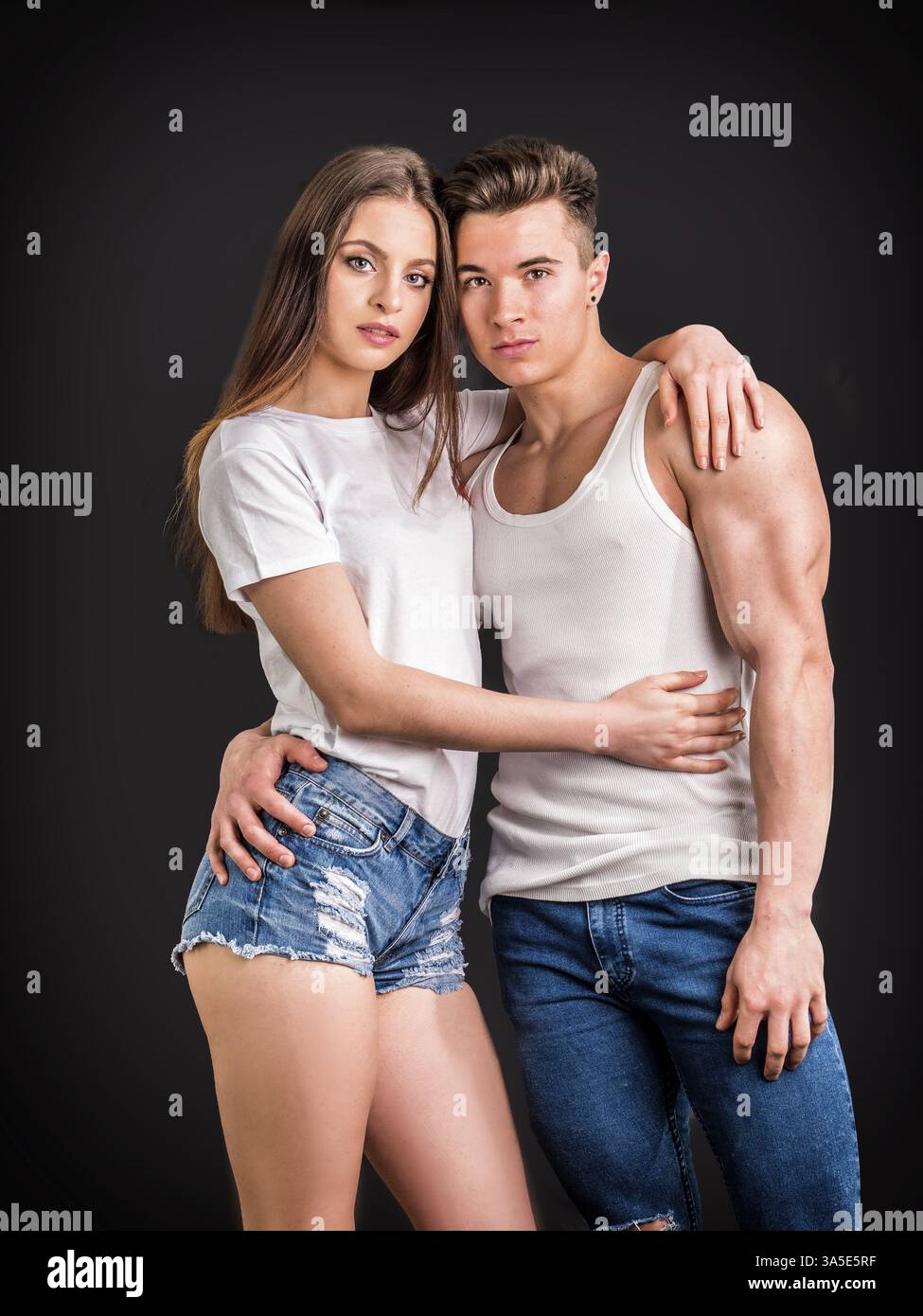 Coppia romantica che abbraccia in studio, l'uomo è muscoloso, la donna indossa pantaloncini in denim e t-shirt bianca, entrambi guardando la fotocamera Foto Stock