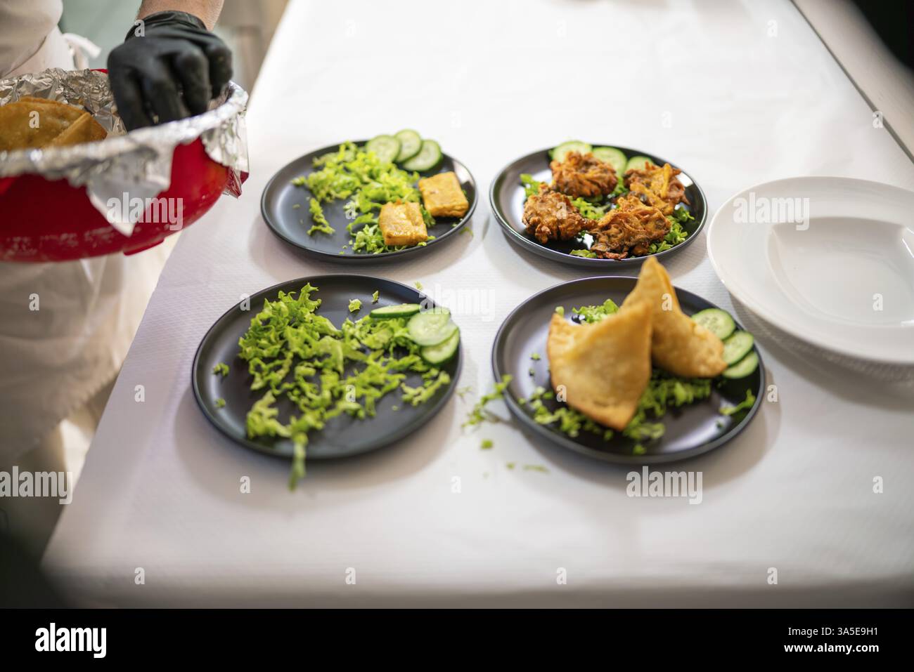 Lo chef indiano indossa guanti neri, posizionando con cura un pezzo di pollo tandoori su un piatto con samosas e insalata, preparando gli ordini da asporto in un riposo Foto Stock