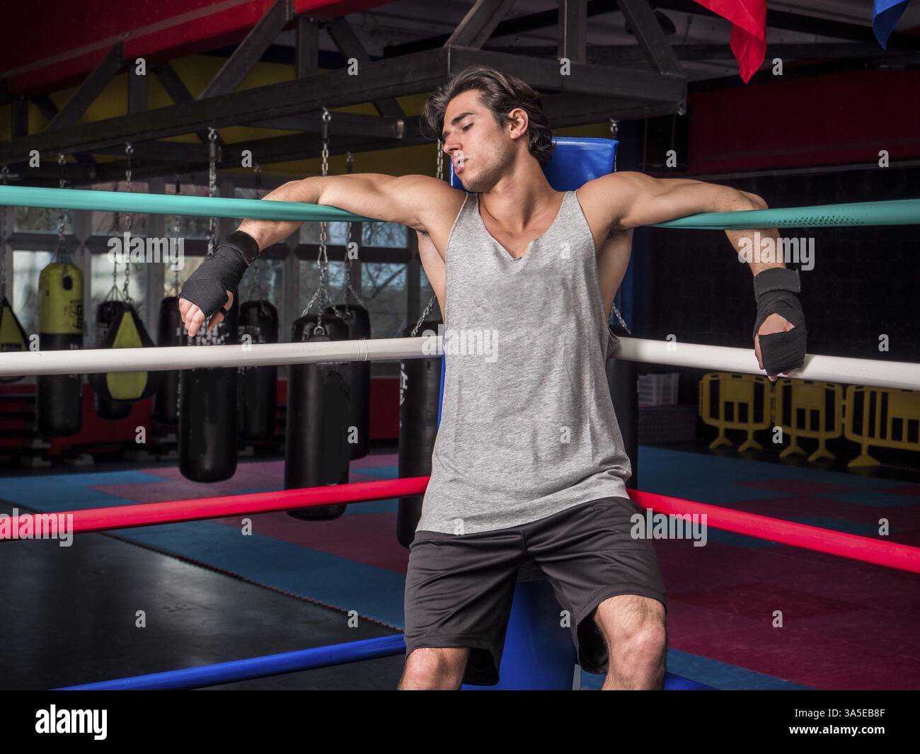 Giovane pugile maschio che si appoggia contro le corde all'angolo dell'anello di boxe, indossando pantaloncini e canotta-top Foto Stock