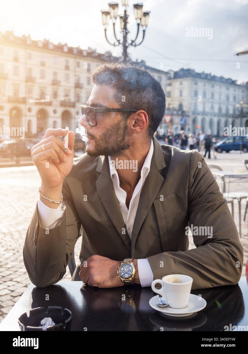 Bel giovane che beve caffè espresso e fuma sigaretta, indossa un elegante cappotto in posa al tavolo all'aperto sullo sfondo urbano Foto Stock