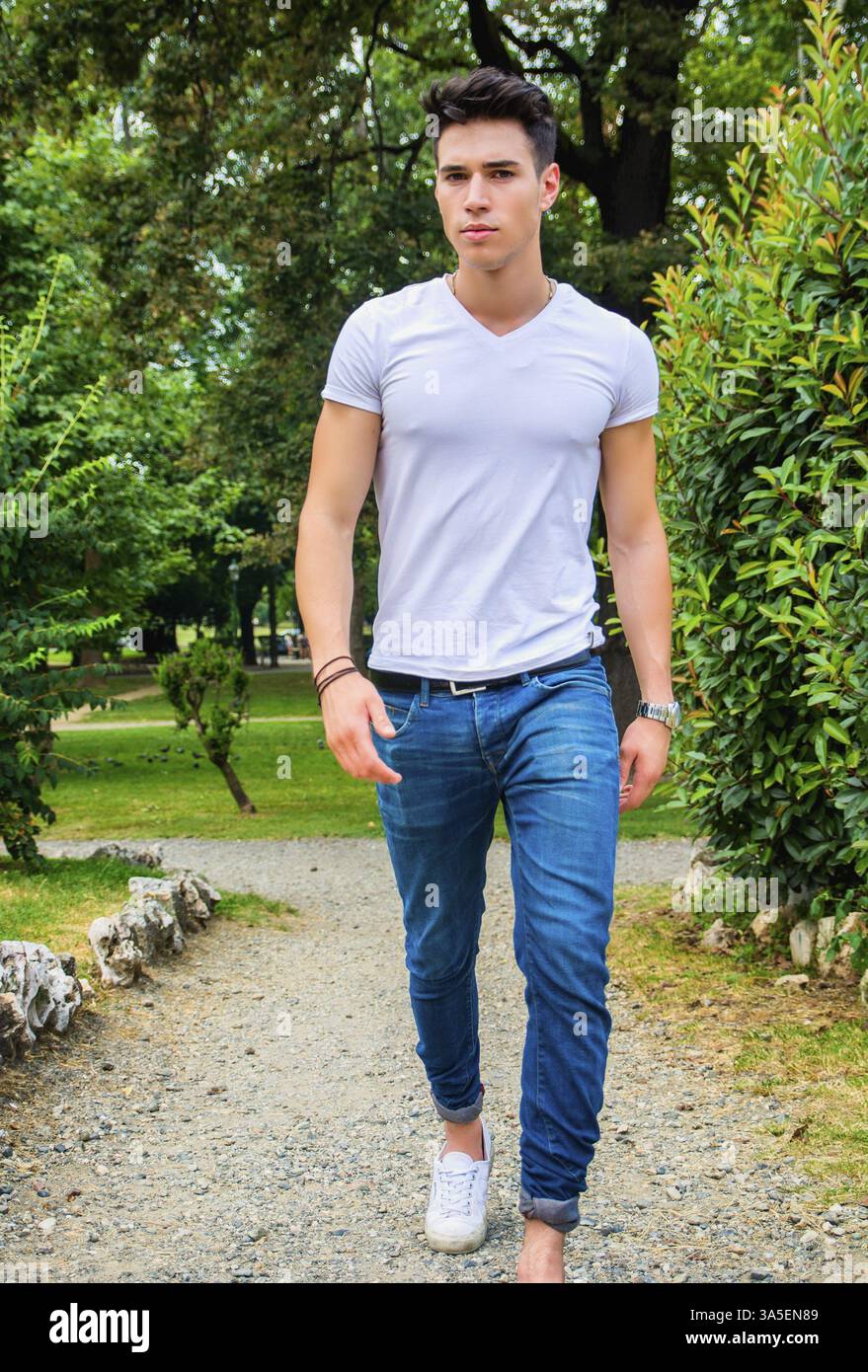 Bel giovane uomo in bianco t-shirt e jeans all' aperto nel parco cittadino, camminando verso la telecamera, gravi Foto Stock