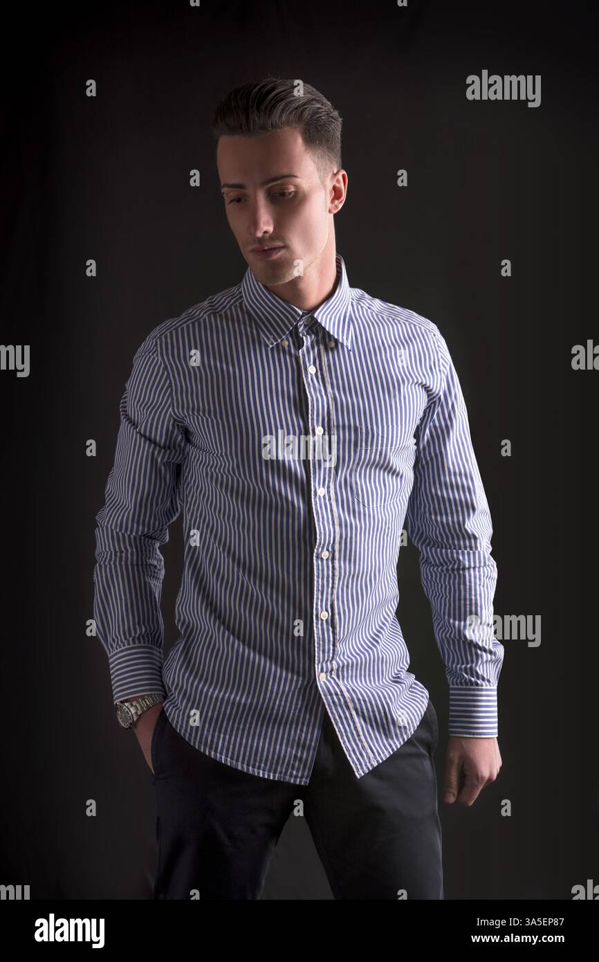 Giovane sorridente in camicia a righe, con lo sguardo rivolto verso il basso su sfondo scuro Foto Stock