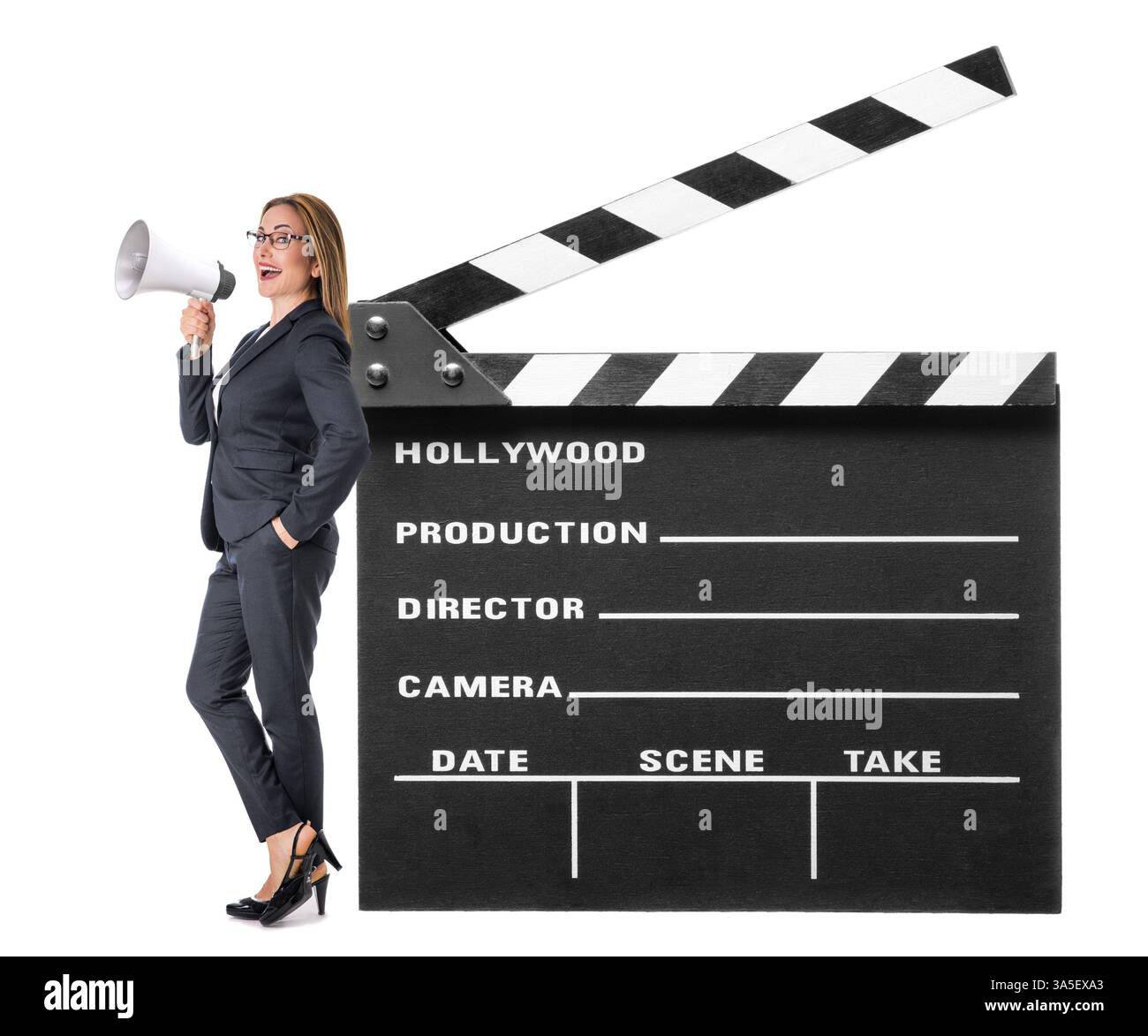 Donna elegante in tuta, regista di film, che parla in megafono mentre si trova vicino a un grande clapperboard su sfondo bianco Foto Stock
