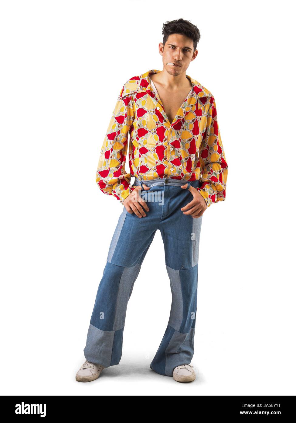 Corpo pieno di elegante giovane modello maschile in camicia alla moda con stampa colorata e jeans svasati che fumano sigaretta e guardano la macchina fotografica, agai isolati Foto Stock