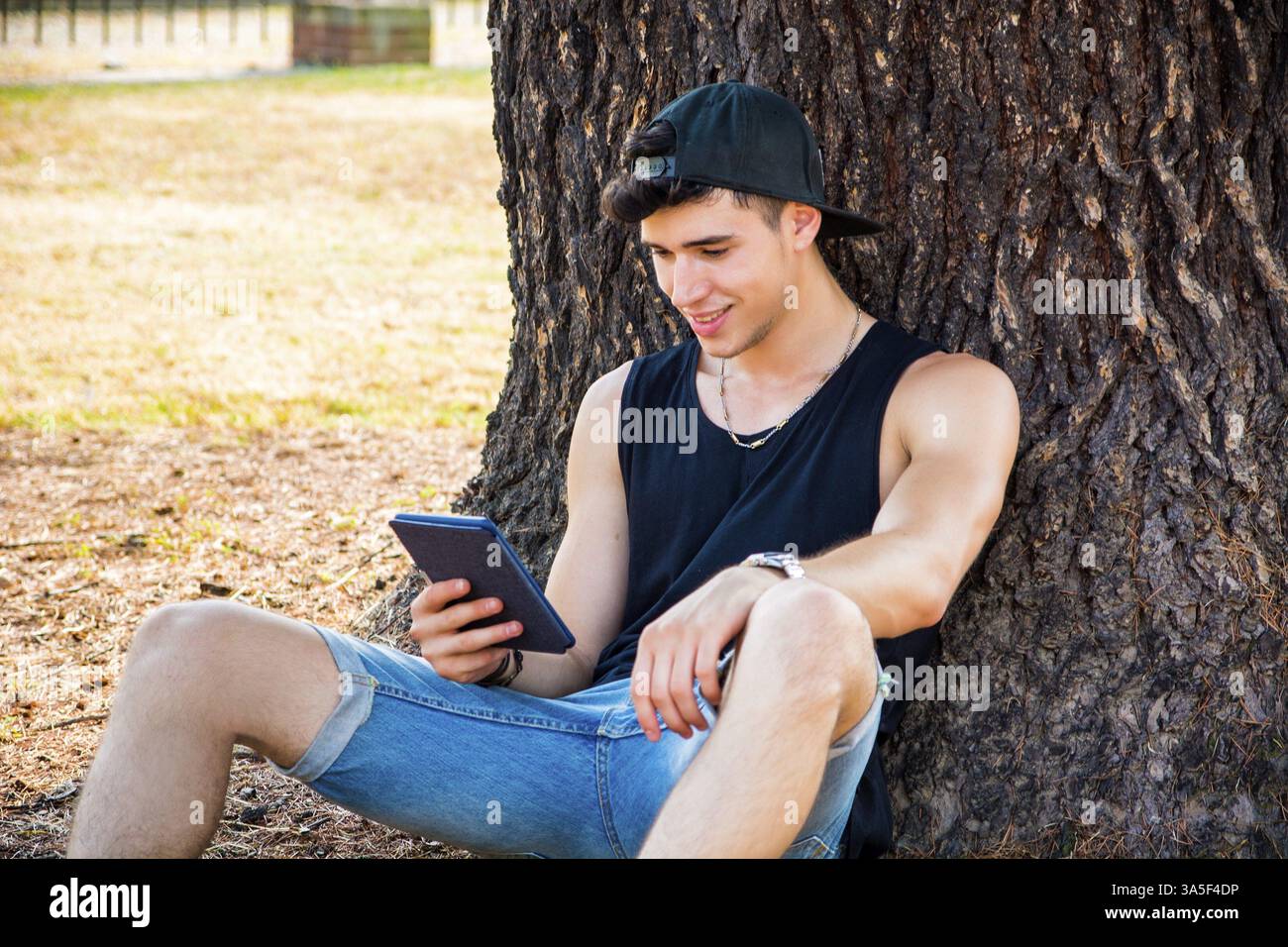 Sorridente Young Handsome Man, leggendo su eBook Reader o tenendo in mano un tablet computer, rilassandosi su un terreno erboso a Park, appoggiandosi sull'albero Foto Stock