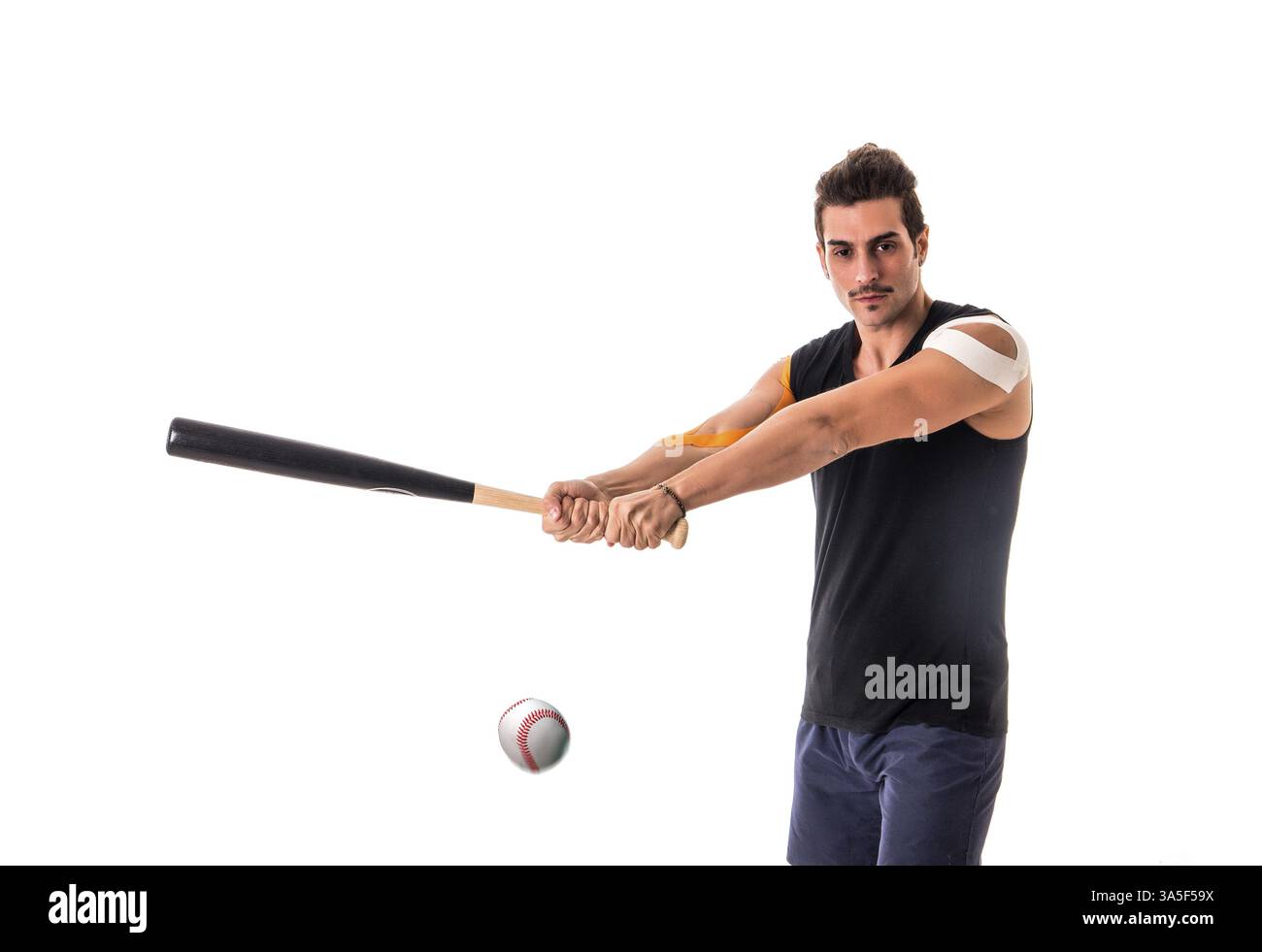 Uomo adulto con nastri Kinesio sulle braccia che reggono la mazza da baseball e sbattono la palla su sfondo bianco Foto Stock