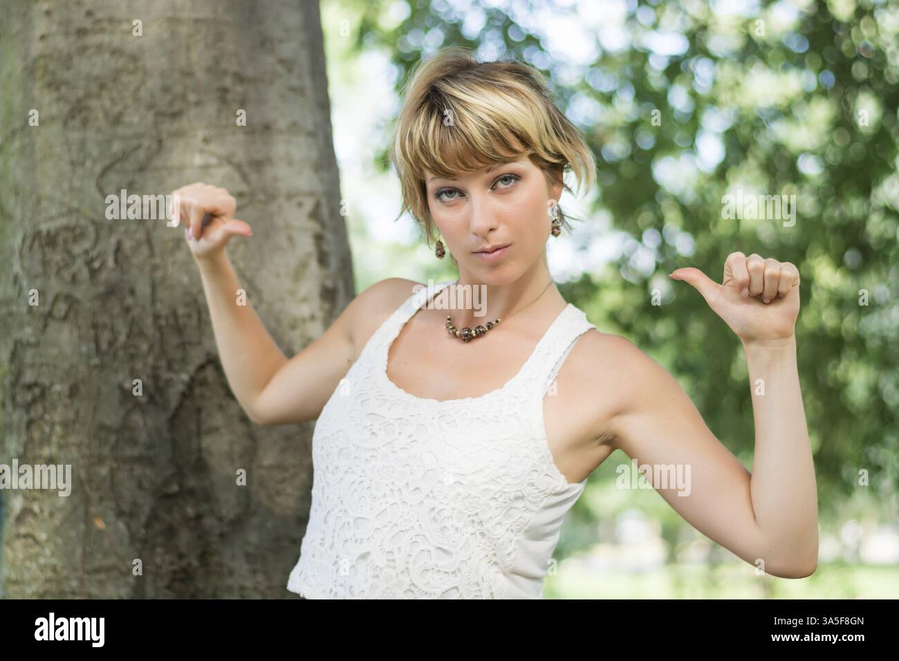 Attraente giovane donna bionda all'aperto che punta le dita verso se stessa, sicura di sé mentre guarda la fotocamera Foto Stock