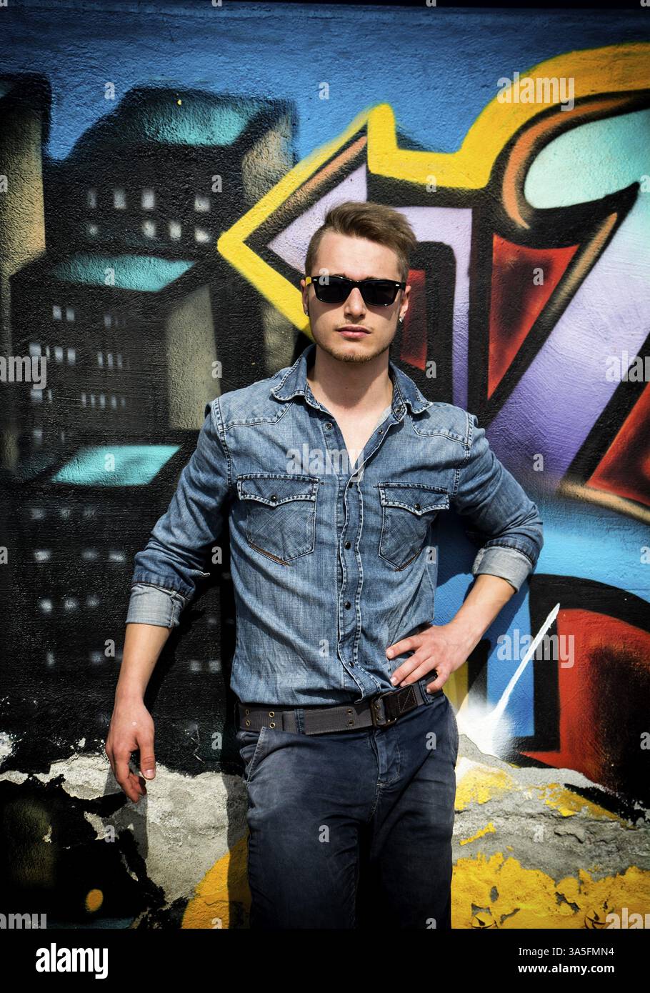 Bella bionda giovane uomo contro il coloratissimo graffito parete, indossare jeans e maglietta jeans Foto Stock