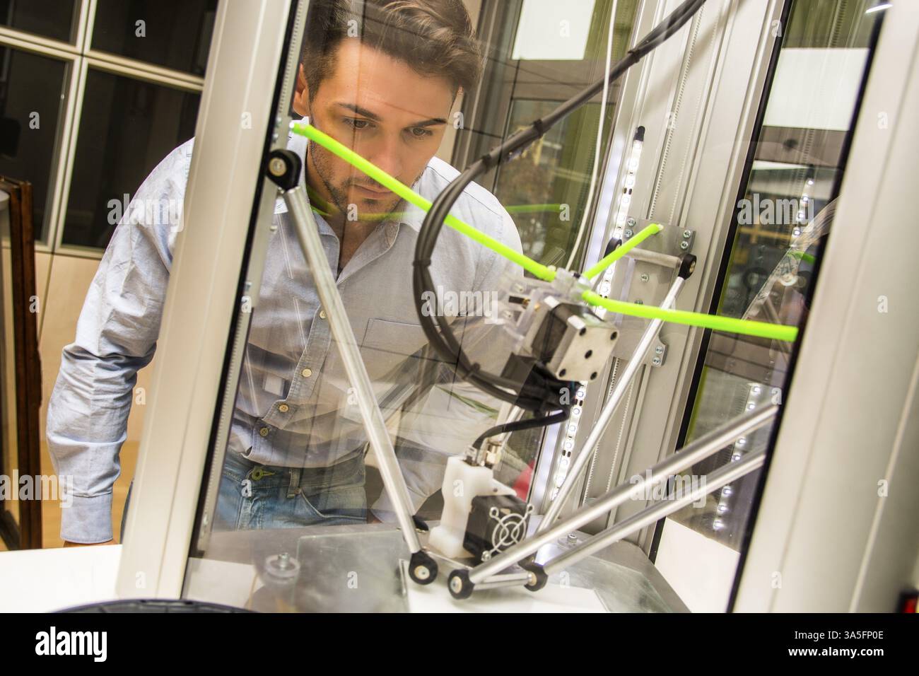 Brunet adulto uomo lavora con stampante 3D, osservando la macchina sul luogo di lavoro Foto Stock