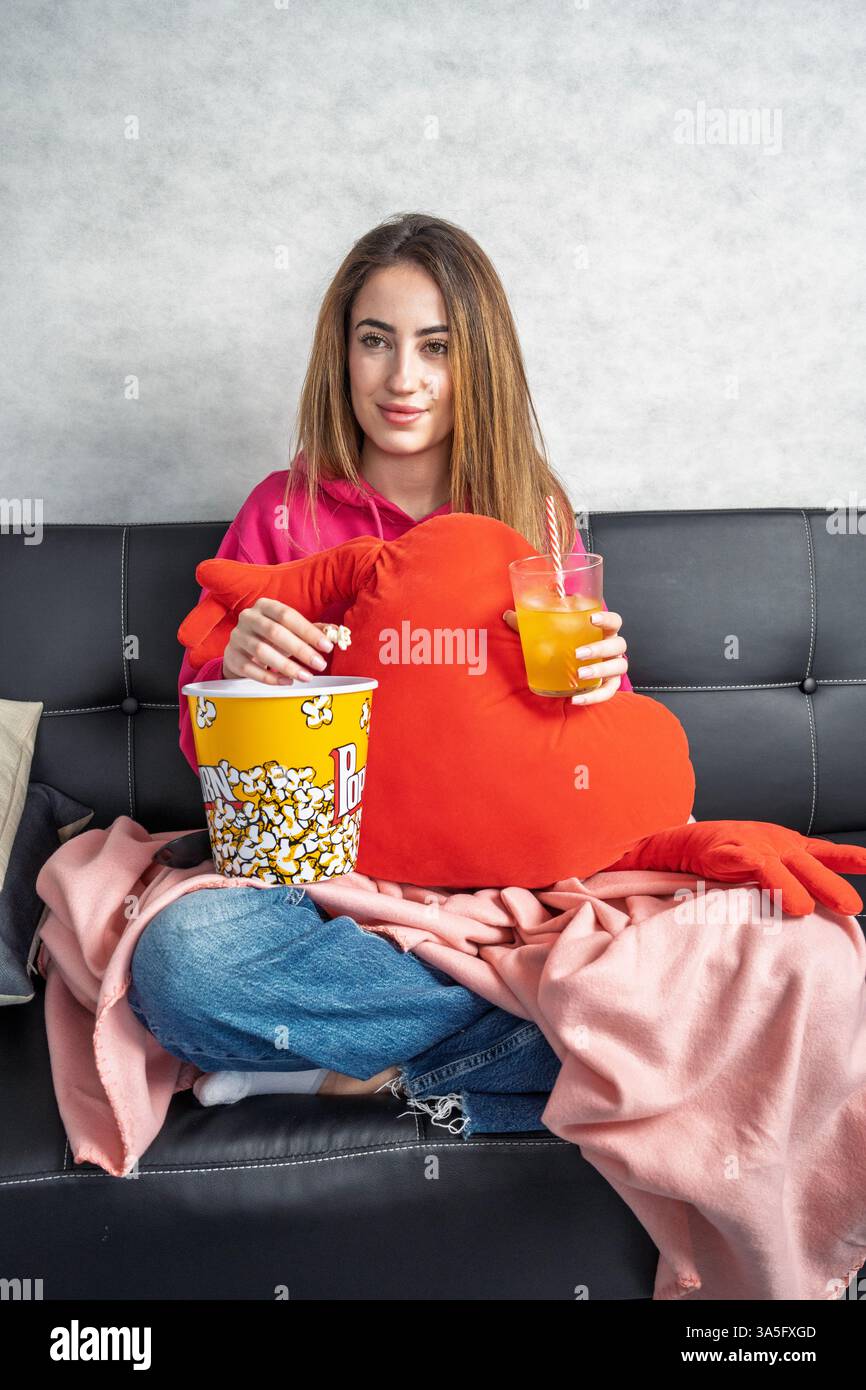 Giovane donna seduta comodamente su un divano, godendosi una serata al cinema con popcorn e succo di frutta Foto Stock