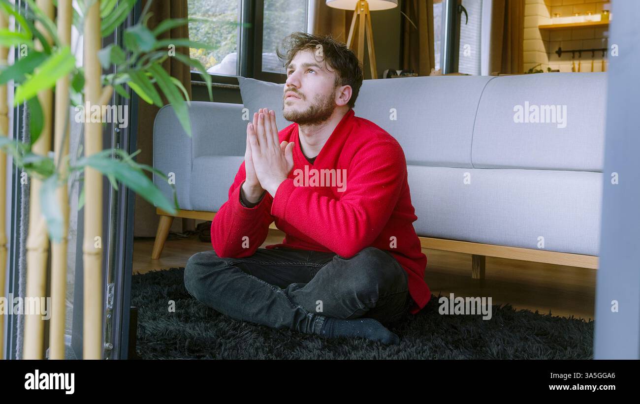 Uomo seduto sul pavimento in posa di preghiera, guardando con attenzione in un ambiente domestico pacifico – mentalità, gratitudine e riflessione spirituale Foto Stock