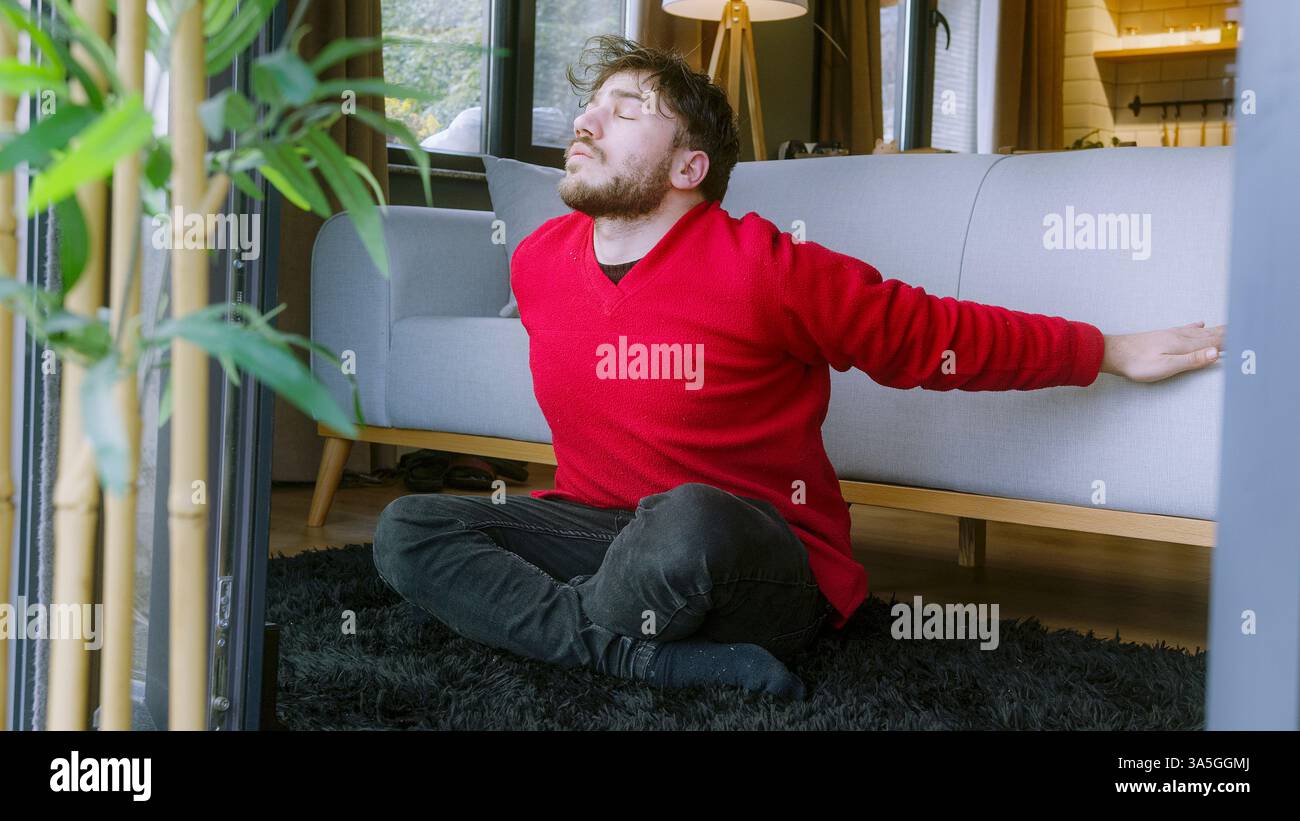 Uomo che allunga le braccia seduto sul pavimento in un ambiente accogliente: Esercizio mattutino e routine di rilassamento Foto Stock