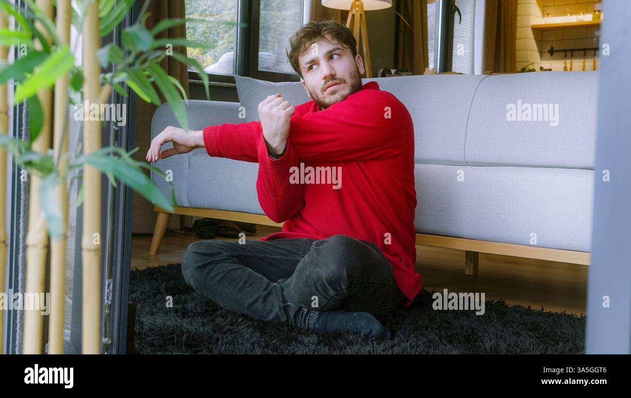 Giovane uomo che allunga le braccia seduto sul pavimento in un ambiente accogliente: Allenamento mattutino e routine di relax Foto Stock