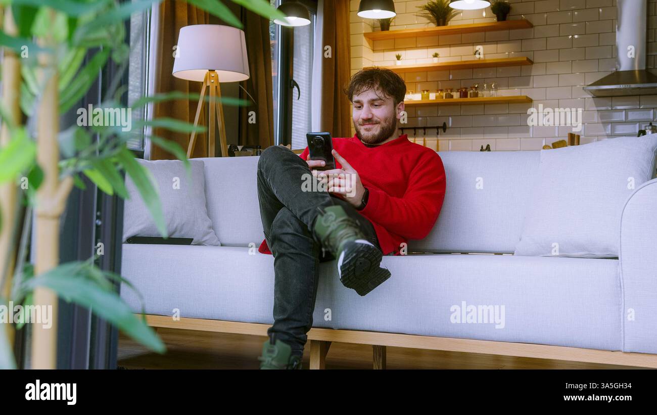 Felice giovane uomo che utilizza lo smartphone mentre si rilassa sul divano nella casa accogliente, sorride e si occupa di comunicazione online Foto Stock