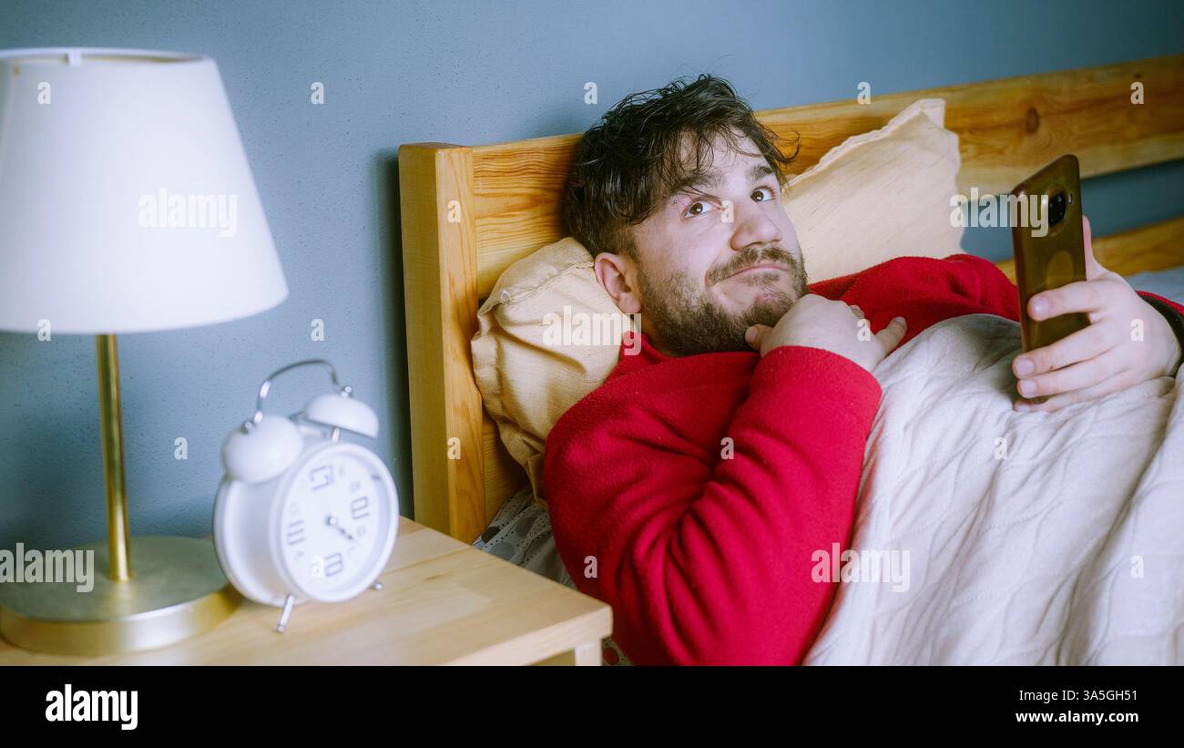 Giovane in maglione rosso sdraiato a letto, con lo smartphone in mano con espressione colpevole, ignorando la sveglia, rimandando in tarda mattinata, distrazione digitale Foto Stock