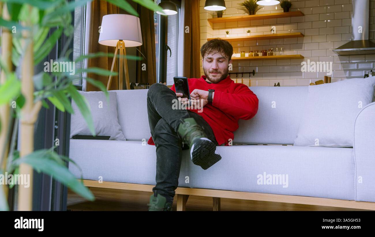 Giovane uomo che utilizza lo smartphone mentre si rilassa sul divano nella casa accogliente, sorride e si occupa di comunicazione online Foto Stock
