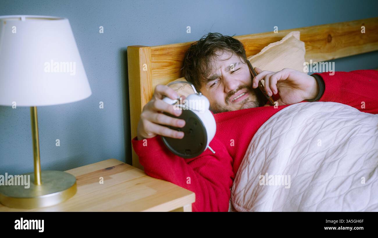 Un giovane a letto che risponde al telefono dal lavoro, tiene la sveglia, non è contento di svegliarsi presto, ha stressato la routine mattutina Foto Stock