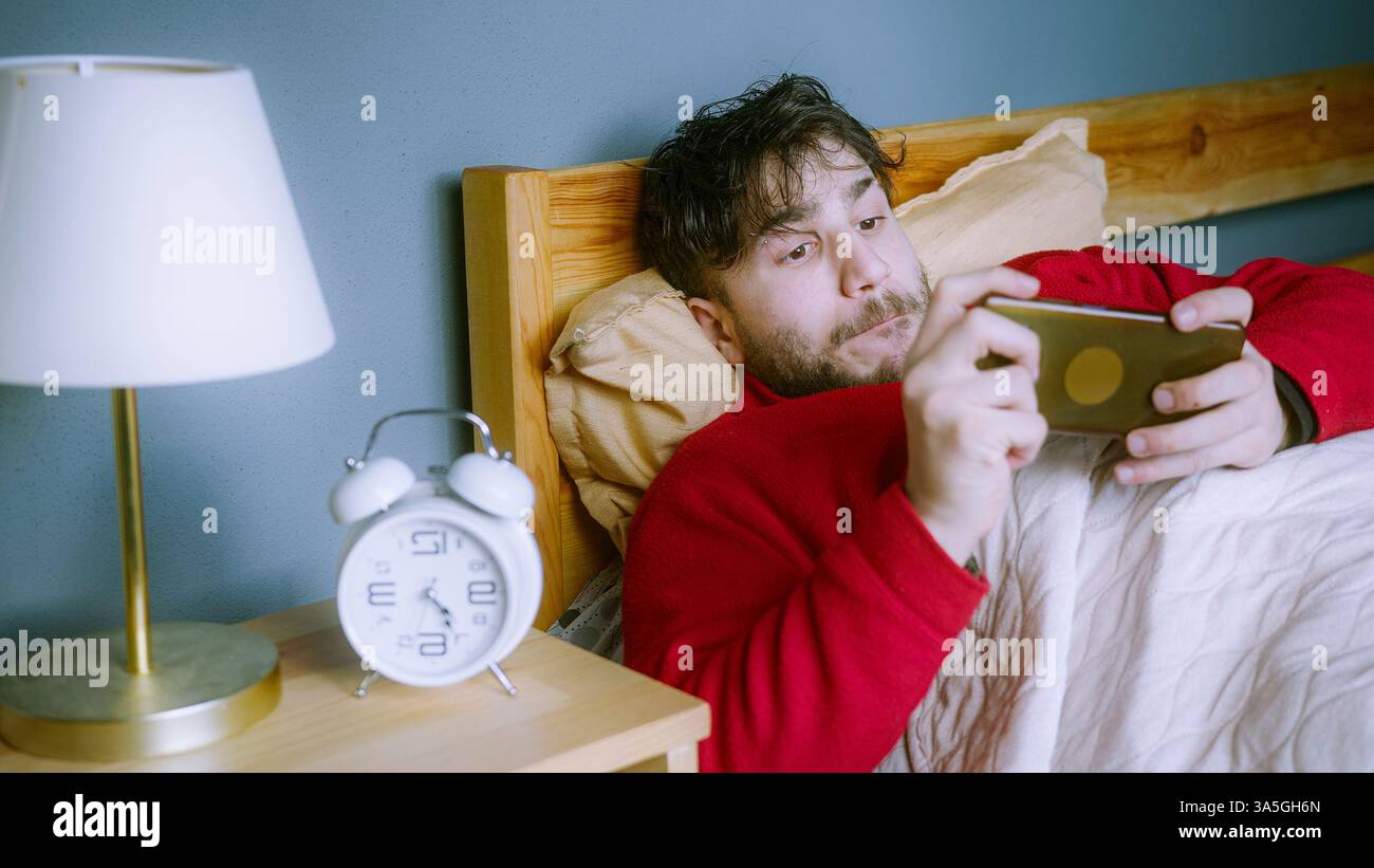 Giovane in maglione rosso sdraiato a letto, gioca a un gioco mobile con una messa a fuoco intensa, ignora la sveglia in camera da letto accogliente, concetto di routine mattutina Foto Stock