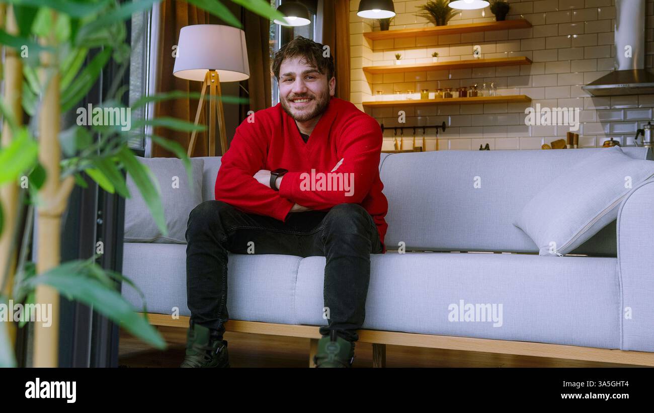 Giovane allegro in un maglione rosso seduto su un divano moderno, sorridente con un'espressione felice e rilassata in un ambiente accogliente ed elegante Foto Stock