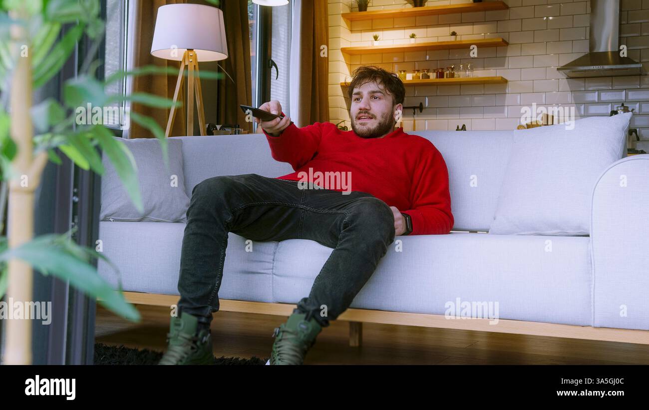 Giovane uomo che guarda la TV a casa, seduto comodamente sul divano con telecomando in mano in un accogliente soggiorno Foto Stock