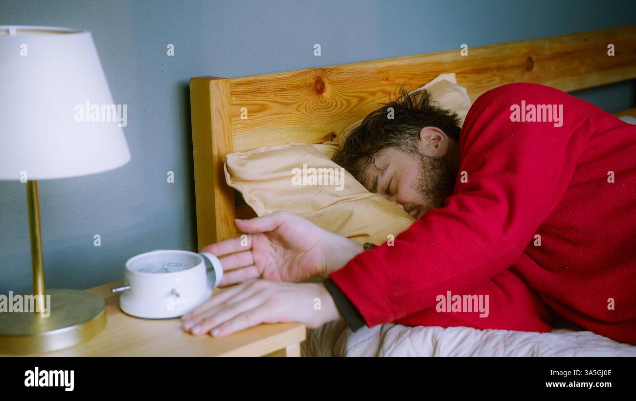 Il giovane lottava per svegliarsi, colpendo Snooze alla sveglia Foto Stock
