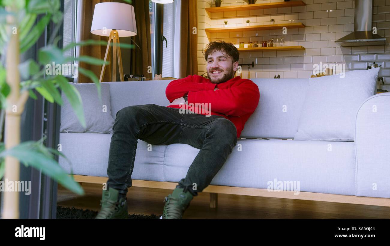 Uomo che guarda la TV a casa, seduto comodamente sul divano con telecomando in mano in un accogliente soggiorno Foto Stock