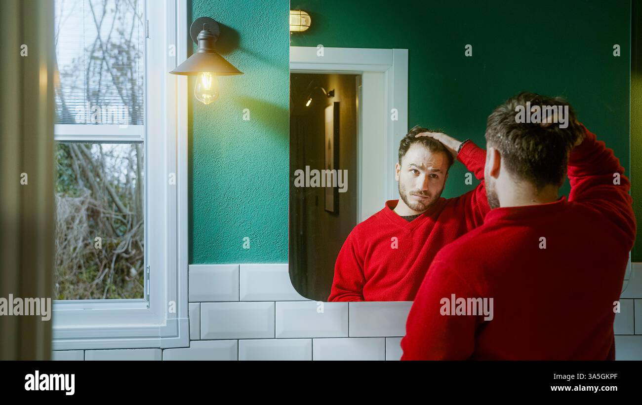Giovane con un maglione rosso che si guarda allo specchio con un'espressione preoccupante, toccando i capelli mentre contempla la perdita di capelli, l'invecchiamento o la sella Foto Stock