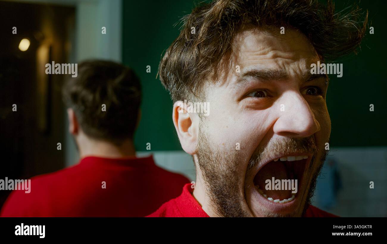 Uomo che urla davanti a uno specchio, esprime frustrazione, rabbia o angoscia emotiva, raffigurando temi di depressione, tumulti interiori e personali Foto Stock