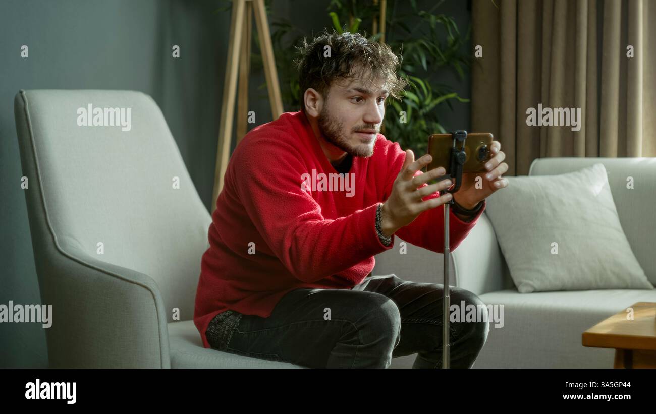 Giovane con un maglione rosso che posiziona il suo smartphone su un cavalletto mentre è seduto in un salotto moderno Foto Stock
