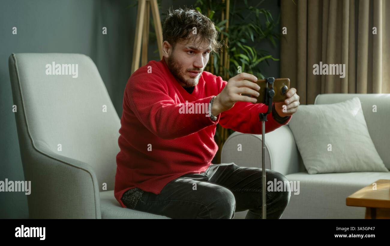 Uomo con un maglione rosso che posiziona il suo smartphone su un cavalletto mentre è seduto in un moderno salotto Foto Stock