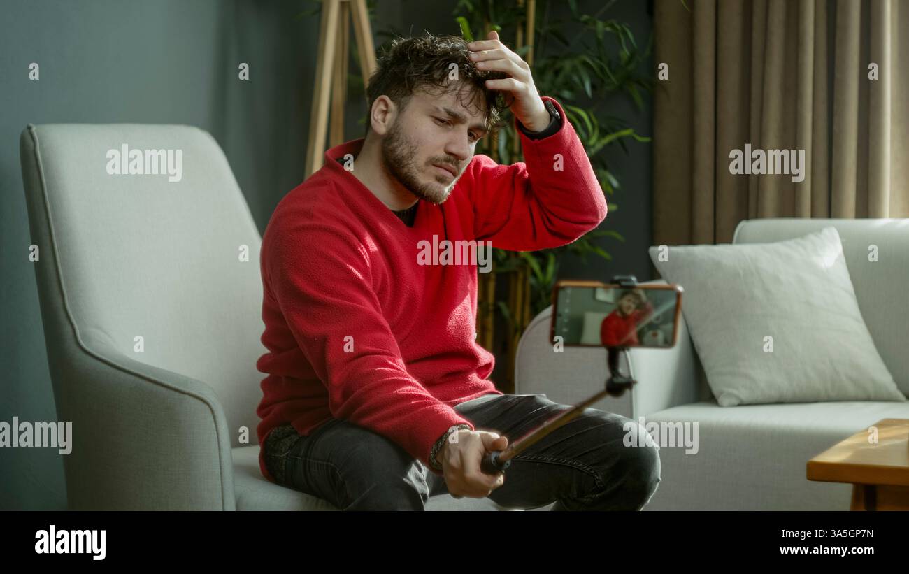 Giovane con un maglione rosso che utilizza un selfie per registrare un video o scattare una foto mentre si siede in un moderno salotto Foto Stock