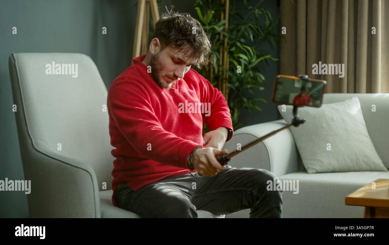Uomo con maglione rosso che utilizza un selfie stick per registrare un video o scattare una foto mentre è seduto in un salotto moderno Foto Stock